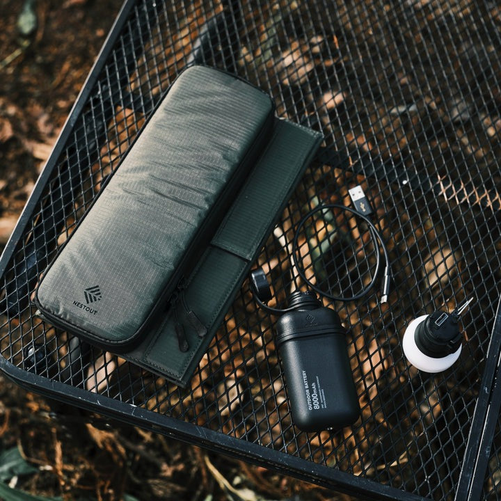 olive green NESTOUT solar panel charger pictured with the NESTOUT waterproof battery pack and lamp accessories | black grate background over fallen leaves in wilderness | survival accessories | lightweight solar panel system for backpacking | essentials for bug out bags